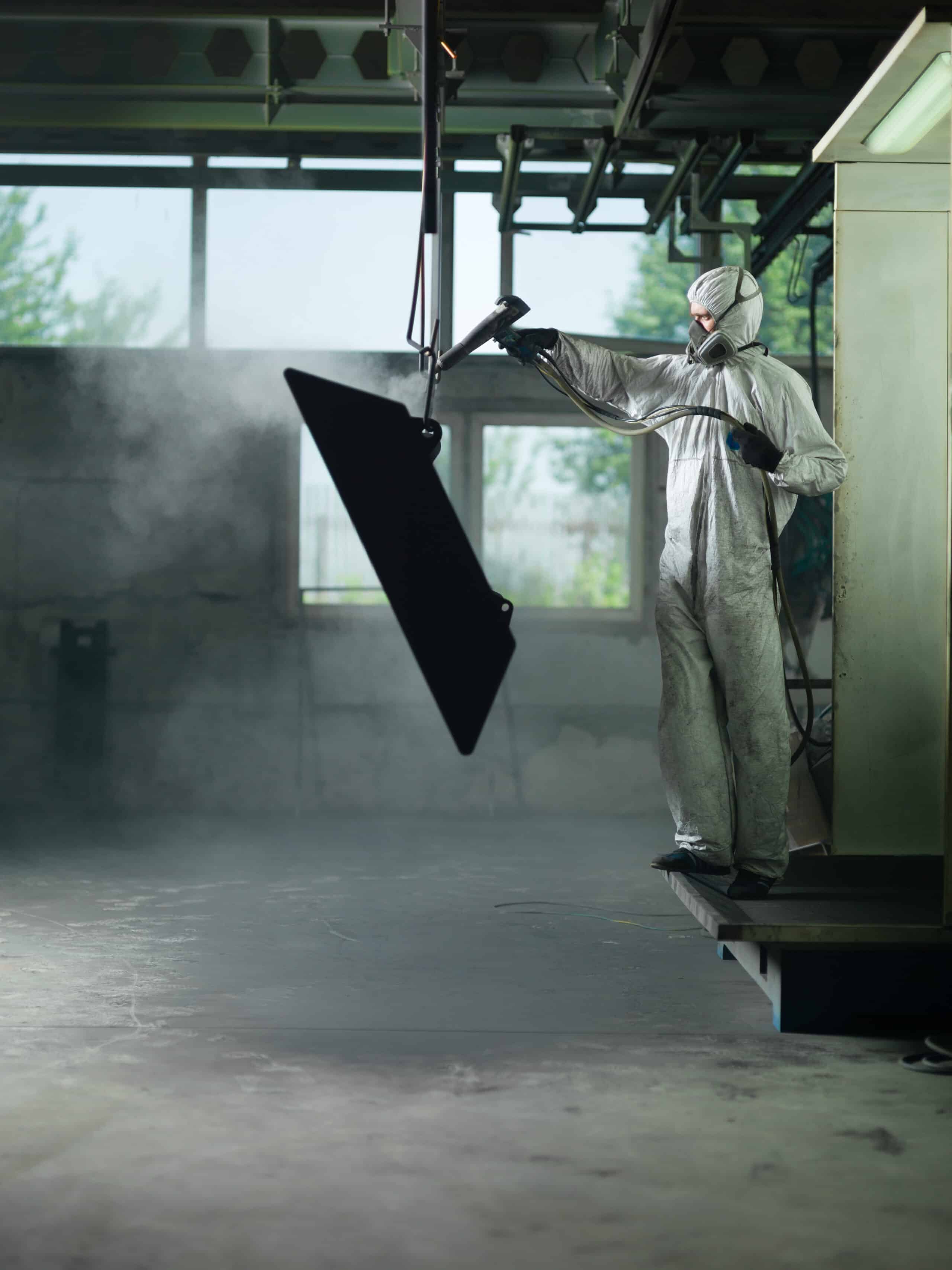 view of a worker wearing a full white protective suit and breathing mask, sand blasting a metal crate hung from a metal beam in the ceiling of an industrial hall